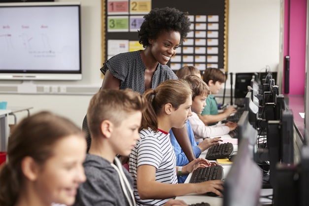 School children sitting in front of computers with a teacher behind them.